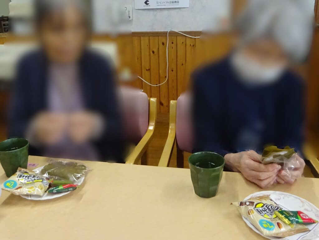食事中の入居者。テーブルに食べ物とグラスが置かれ、複数の人物が見える。