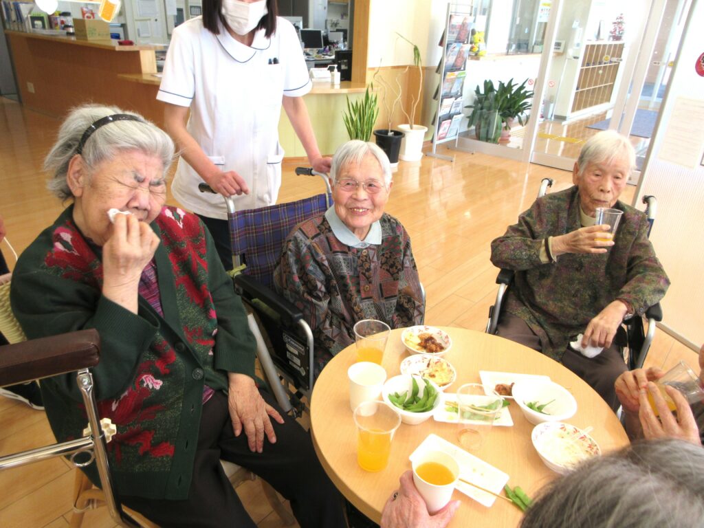 介護施設での食事風景。入居者3名がテーブルで食事をしており、スタッフが見守っている。